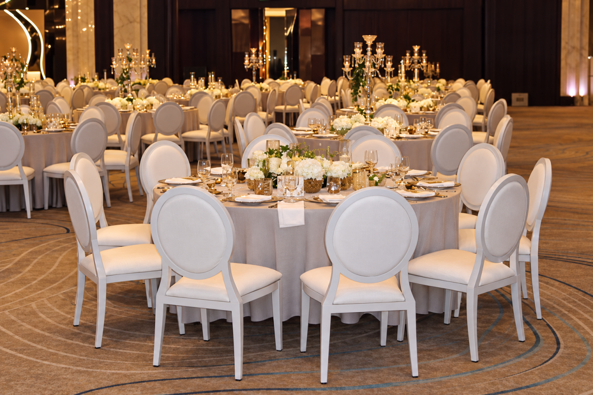 White Dior dining chairs arranged around round banquet tables with dinnerware and low white floral centrepieces in Dubai.